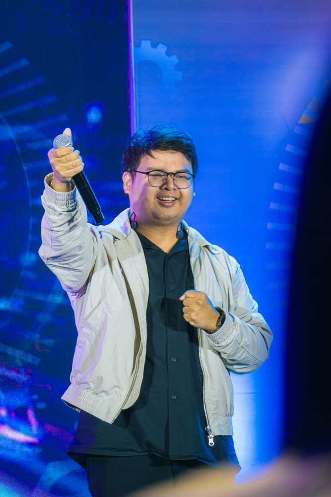 A speaker in a light jacket smiling and pointing his microphone toward the audience on a blue-lit stage.