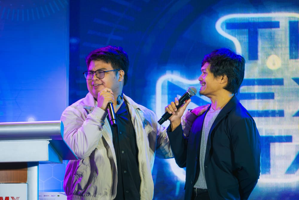 Two speakers sharing microphones and laughing together on stage in front of a large screen.