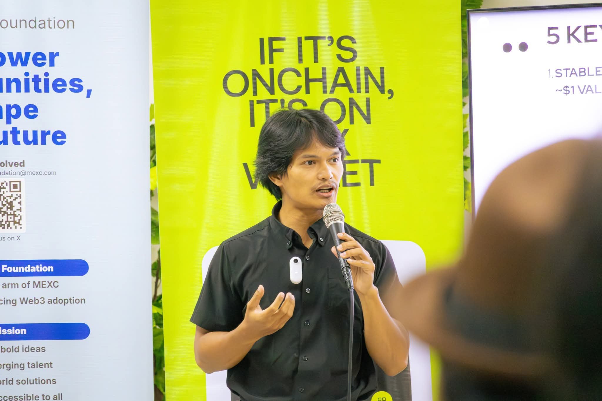 Jops speaking at a Web3 community event, holding a microphone in front of a branded stage backdrop.